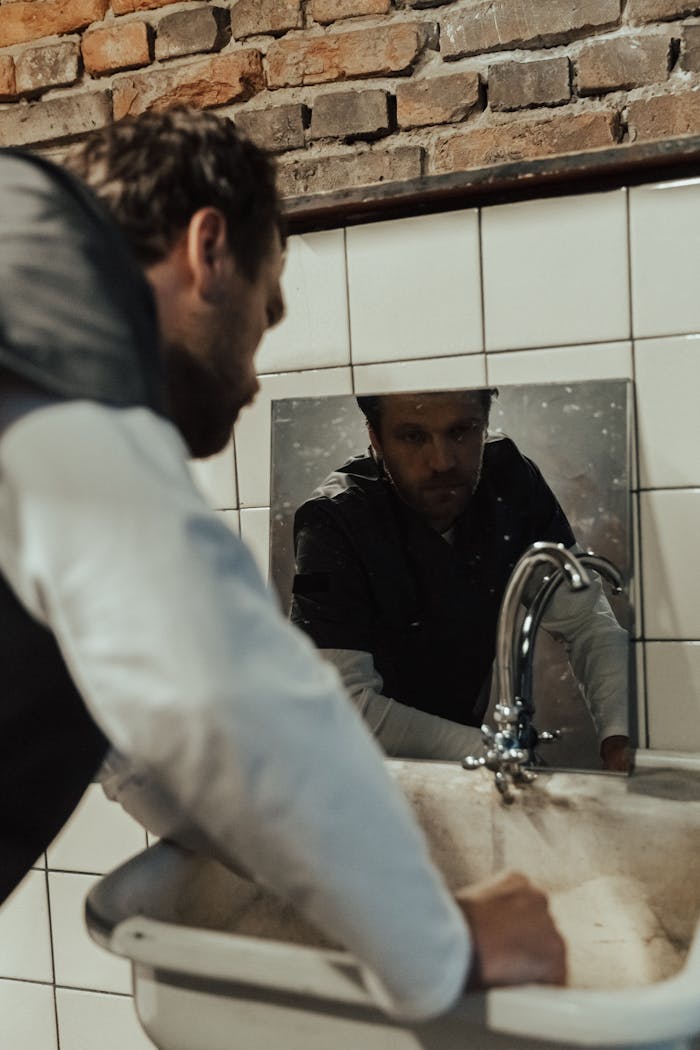 Man staring at mirror above an old bathroom sink in rustic setting, conveying introspective mood.